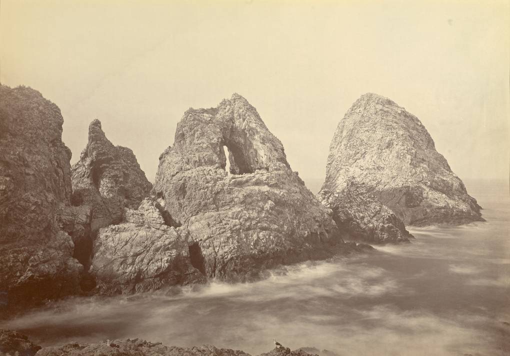 A sepia toned photo of tall rocky land masses surrounded by foamy sea water.