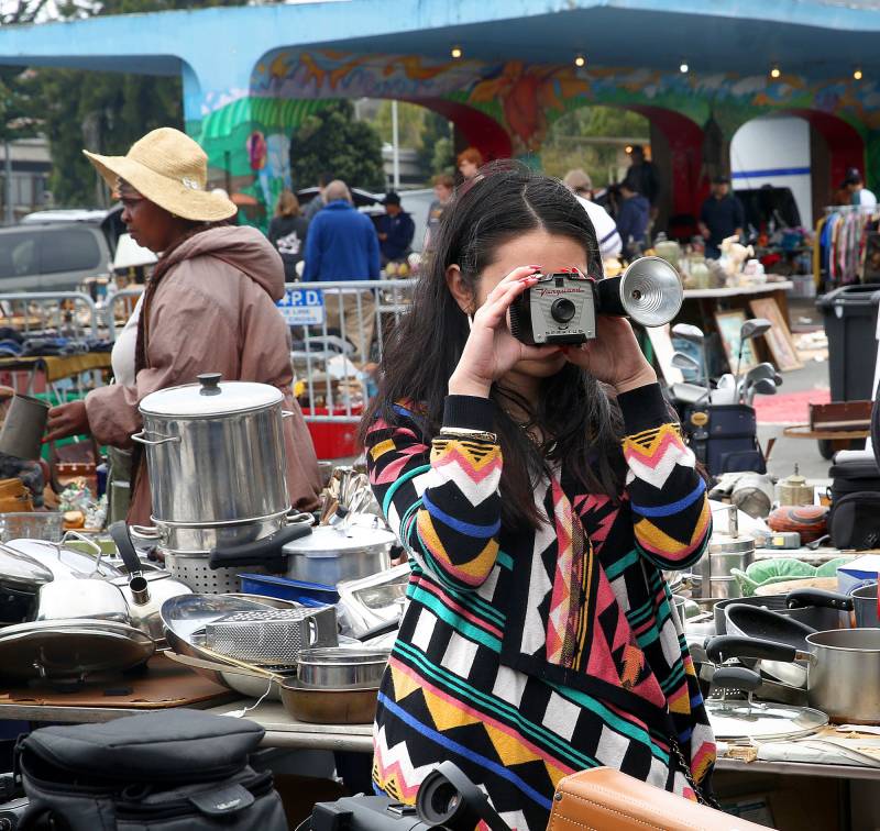 Person holds a vintage camera to their face surrounded by housewares and other used objects