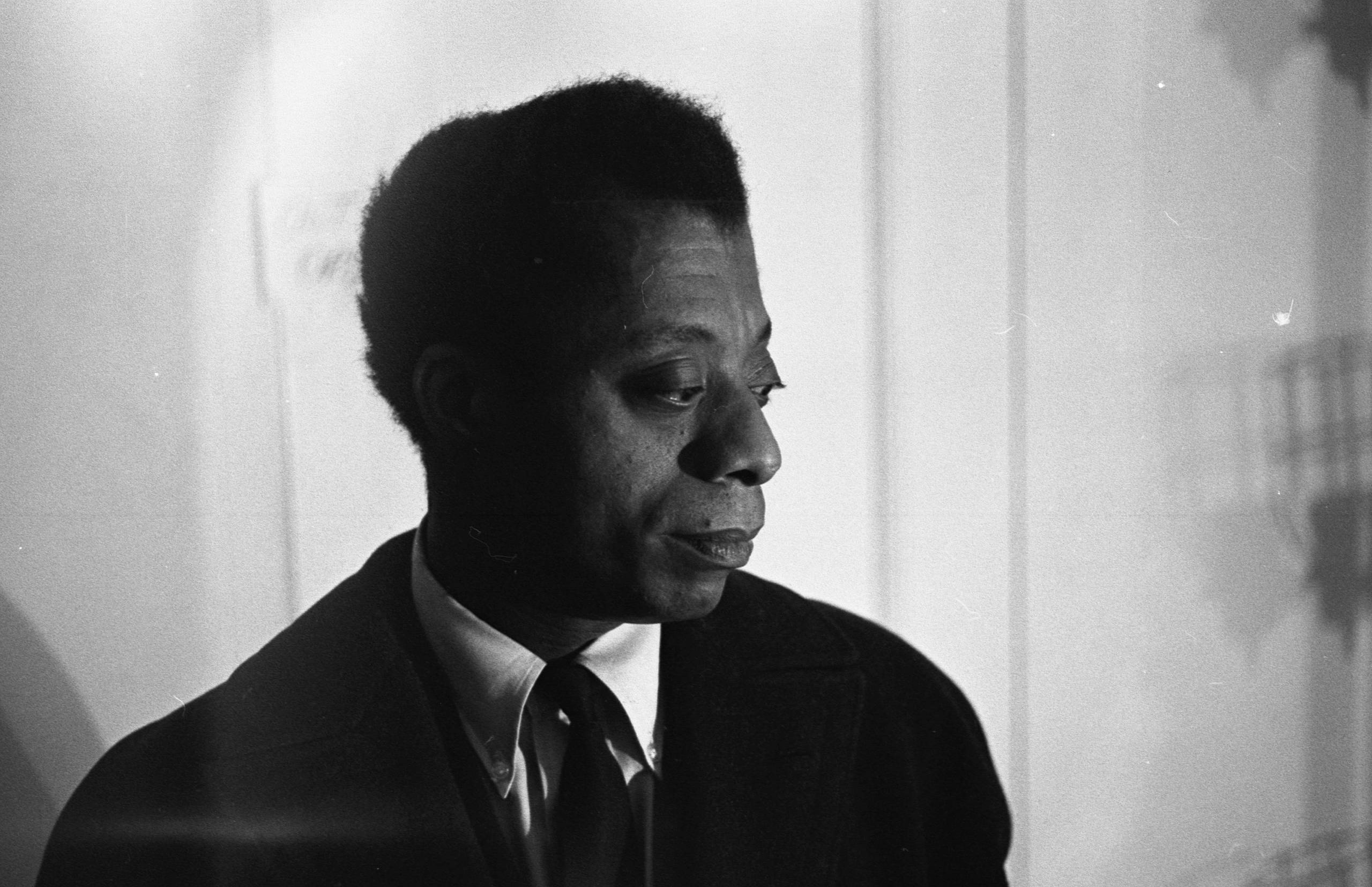 A Black man wearing a suit stands in front of a white wall with his head turned to one side.