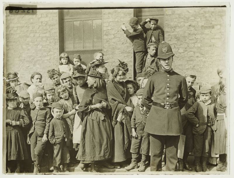 An old fashioned policeman stands in front of a group of scrappy-looking children of various ages.