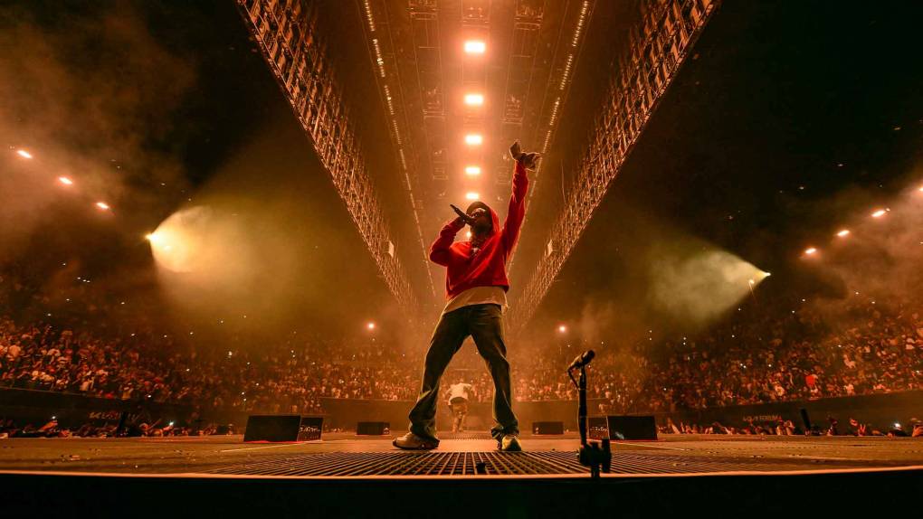A Black man in a red hoodie raps on the mic and holds one finger aloft while an arena ceiling bathed in golden light is in the background