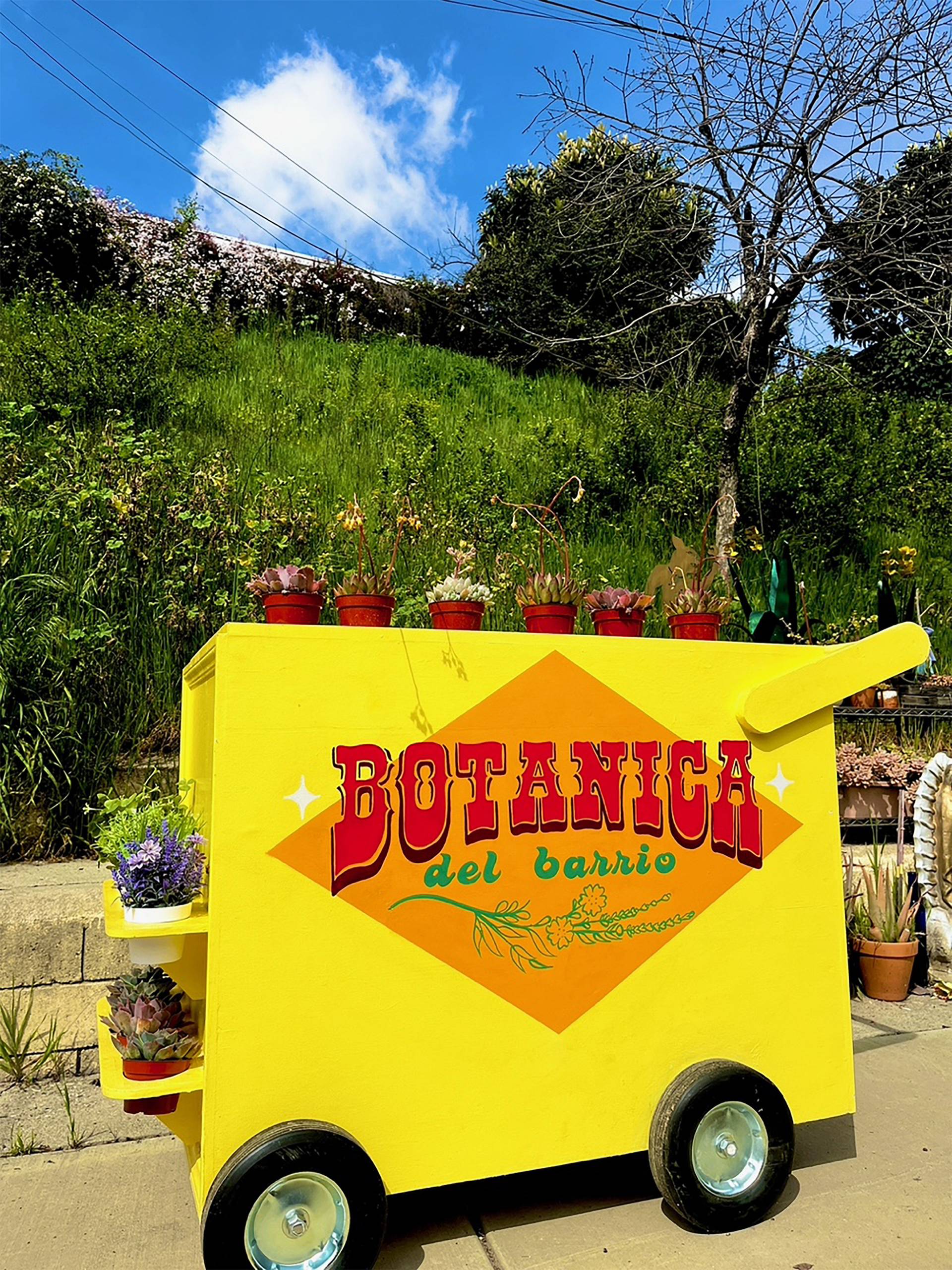 a yellow push cart labeled as Botanica del Barrio is displayed outdoors near a garden