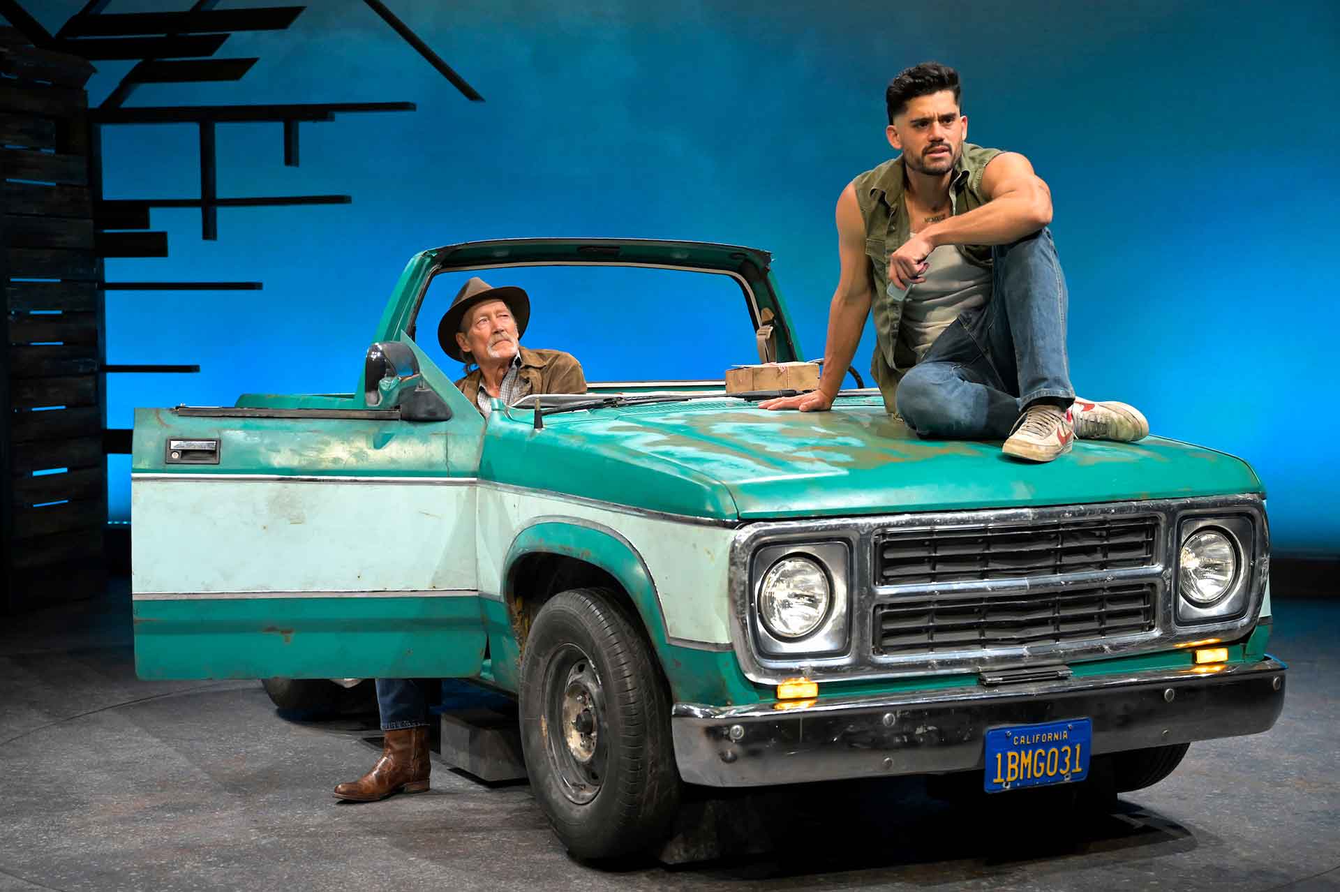 An older man sits inside a truck cab while a younger man in a sleeveless shirt and jeans sits on the hood.
