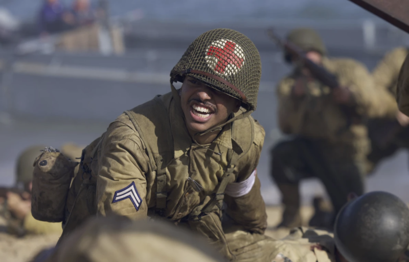 A Black man wearing World War II army fatiques and a medic helmet crouched over a body on a chaotic beach.