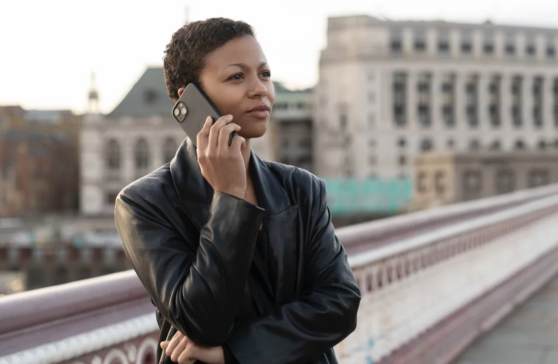 A Black woman with very short hair stands on a bridge, holding a cell phone to her ear.