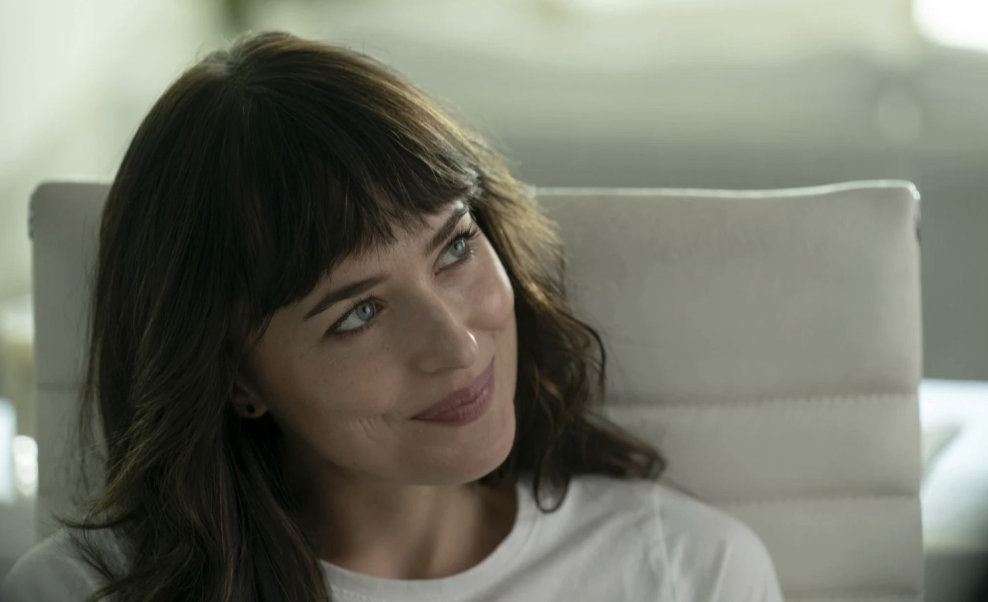 A white woman with long brown hair gazes up at someone and smiles, while seated in a white chair.