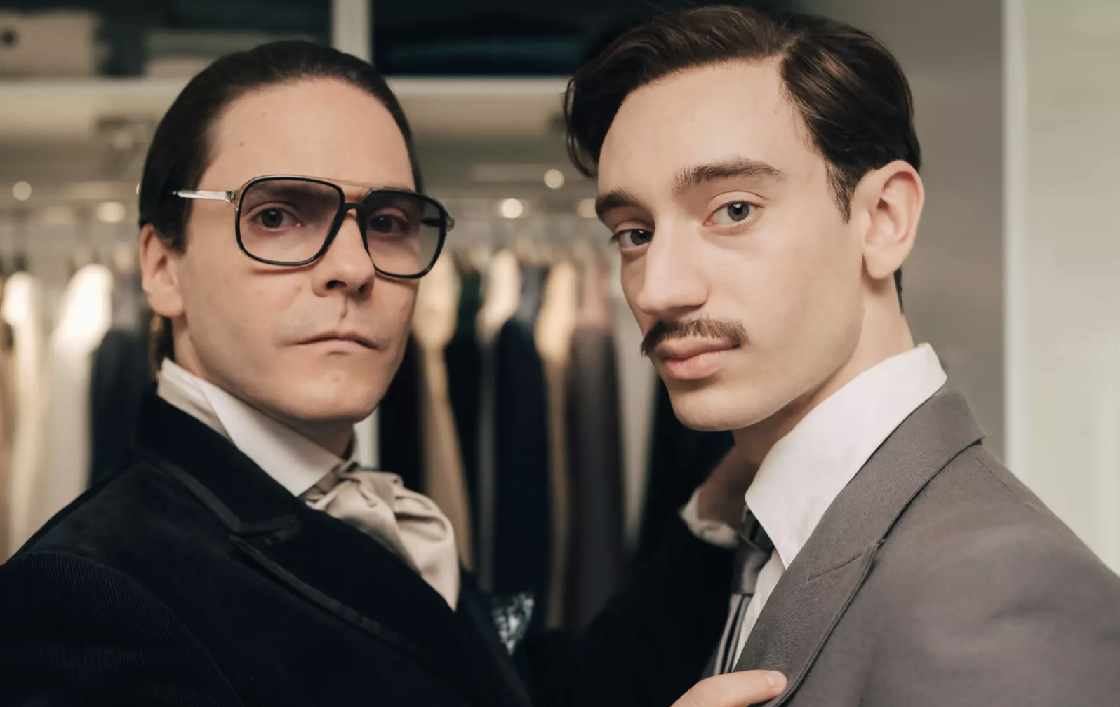 Two men stand with their bodies close together in front of a clothes rack. They are both looking towards the camera.