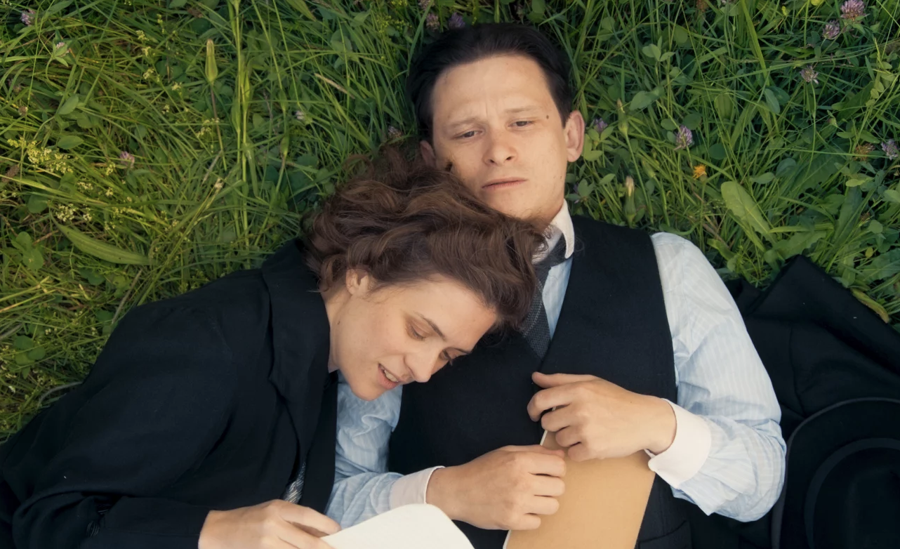 A man lies in the grass, while a woman lies partially on his chest, happily reading a letter.