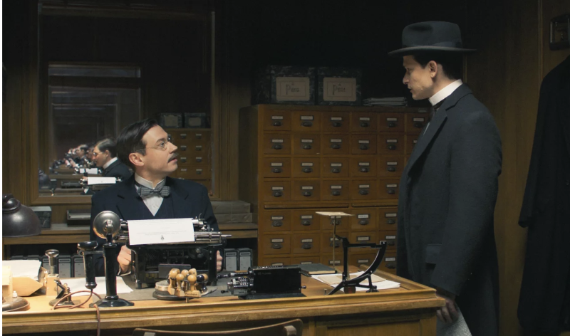 A man stands next to the desk of another man in turn of the century Europe.