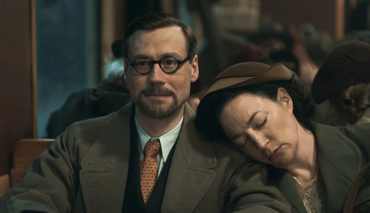 A man wearing a suit and spectacles sits with a woman leaning on his shoulder who has fallen asleep.