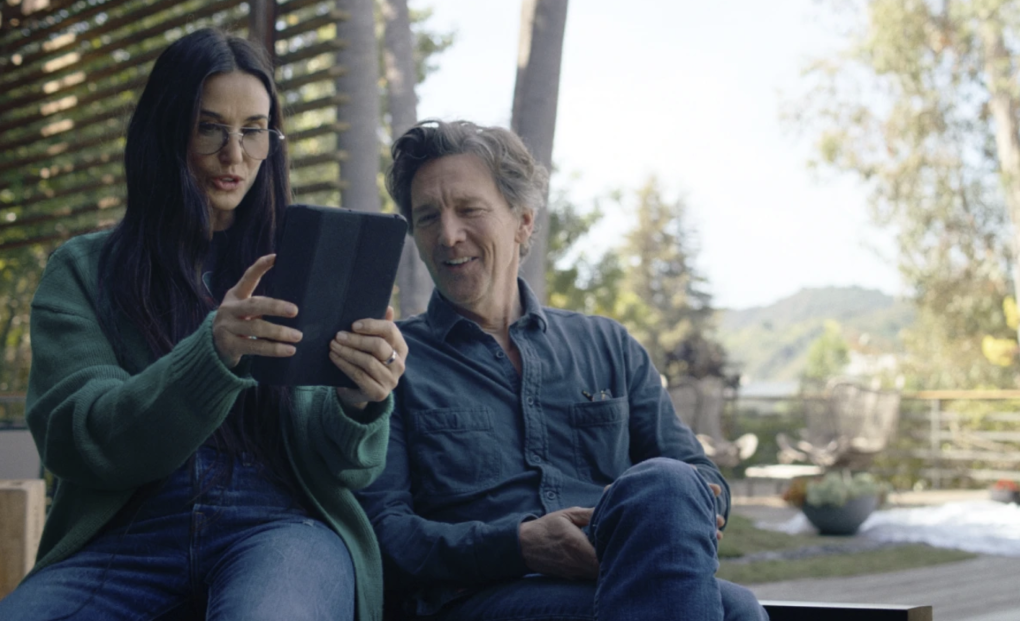 A woman and man sit together on a porch staring at the screen of an iPad, visibly amused.