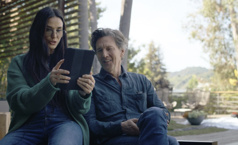 A woman and man sit together on a porch staring at the screen of an iPad, visibly amused.