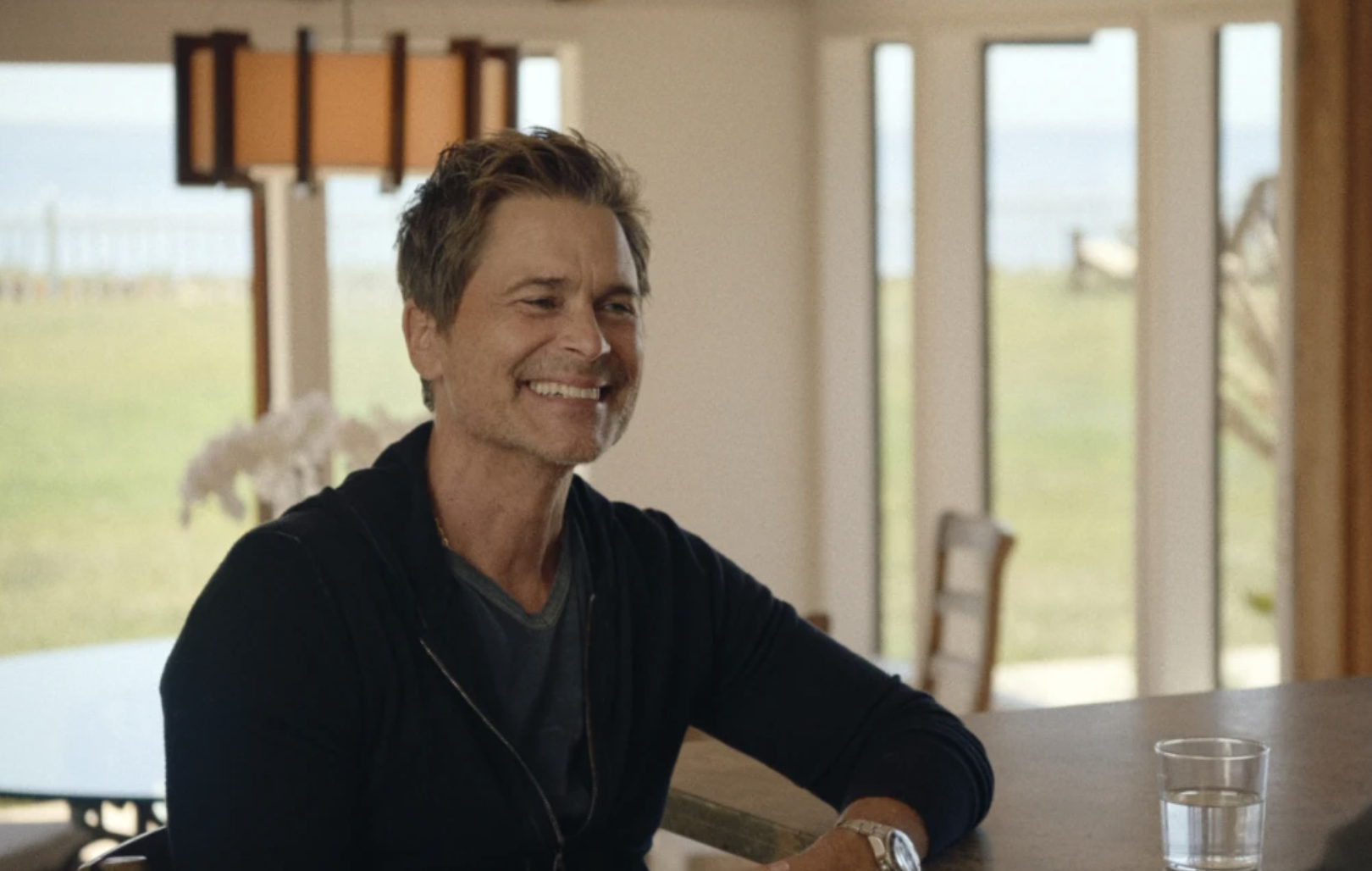 A handsome middle-aged man sits at a kitchen counter, laughing warmly.