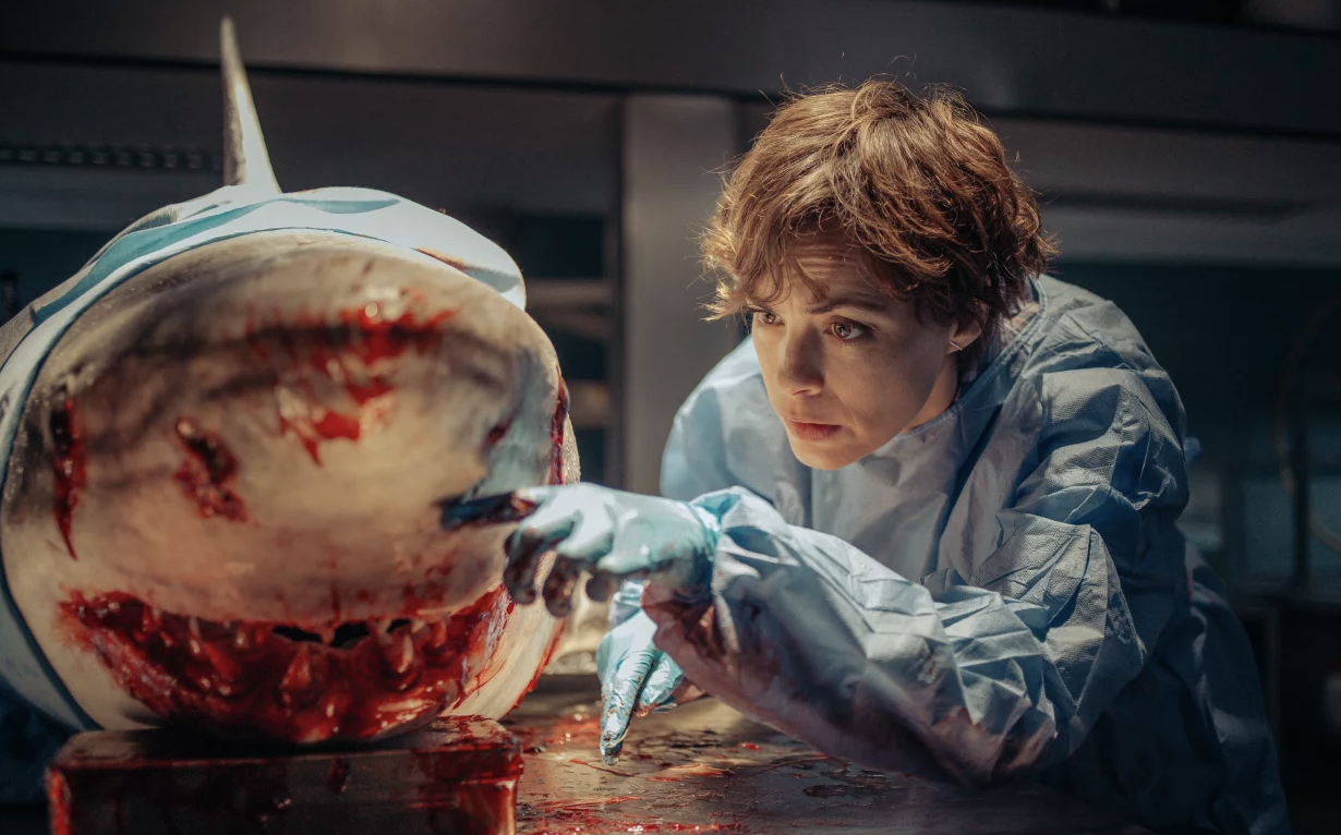 A woman in scrubs examines a dead, blood-covered shark.