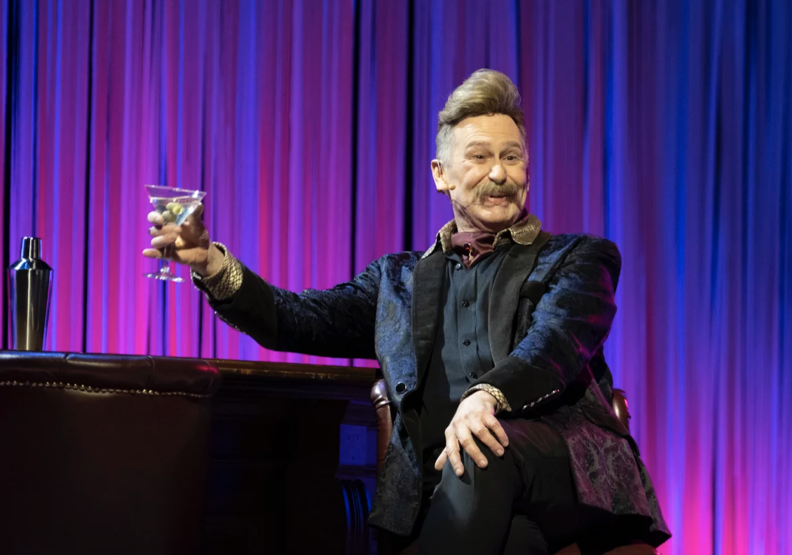 A dapper man sits in front of a purple curtain, smiling and raising a cocktail.