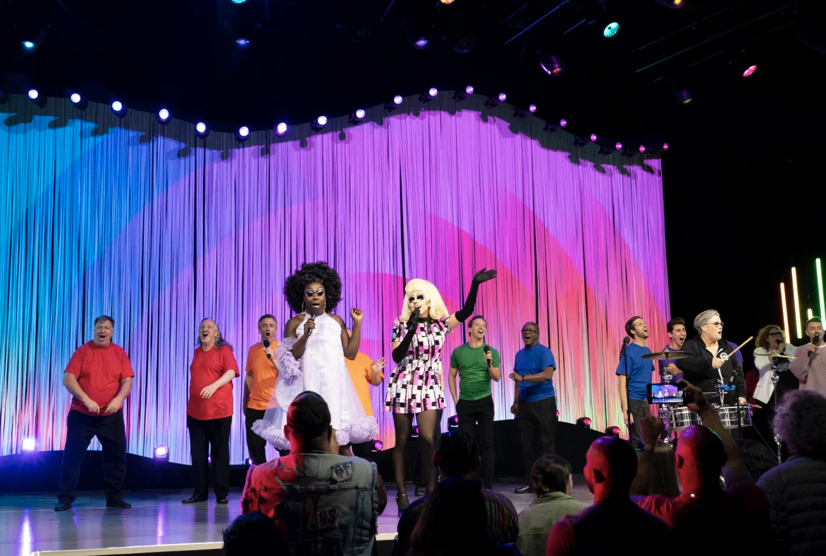 Two drag queens perform at the front of a stage, while a chorus sings behind them.