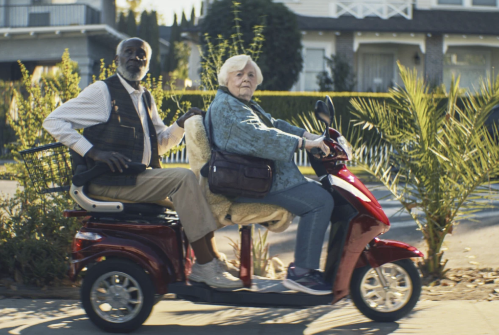 A senior white lady with white hair rides on a mobility scooter with a snappily dressed senior Black man riding on the back.
