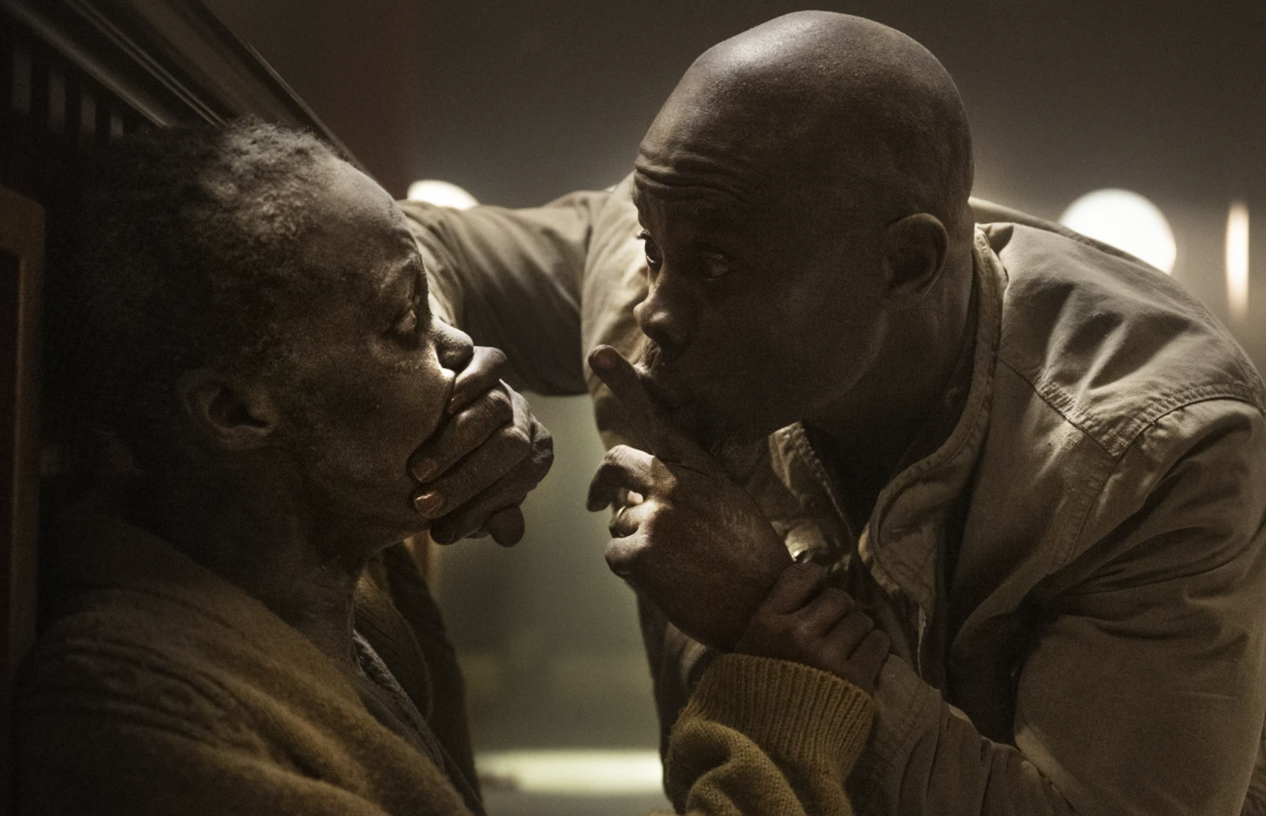 A Black man crouches down with his finger to his lips. Hi hand is covering the mouth of a Black woman seated in front of him. They are both covered in white dust.