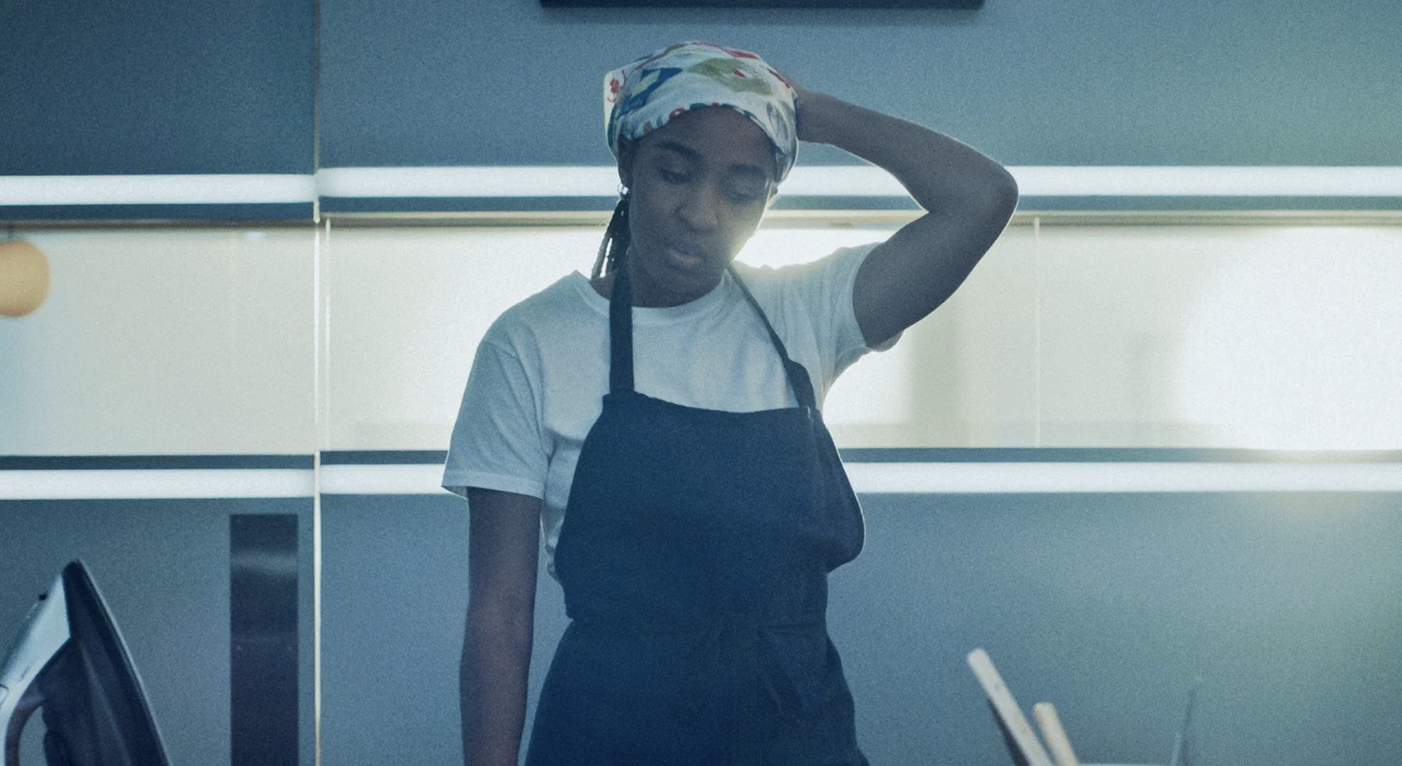 A Black woman stands in a kitchen looking stressed. She is wearing a white t-shirt and black apron.