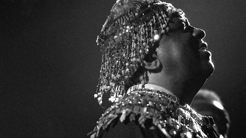 A Black man in a glittery headpiece and shoulder sash tilts his head back, facing right, seen in profile