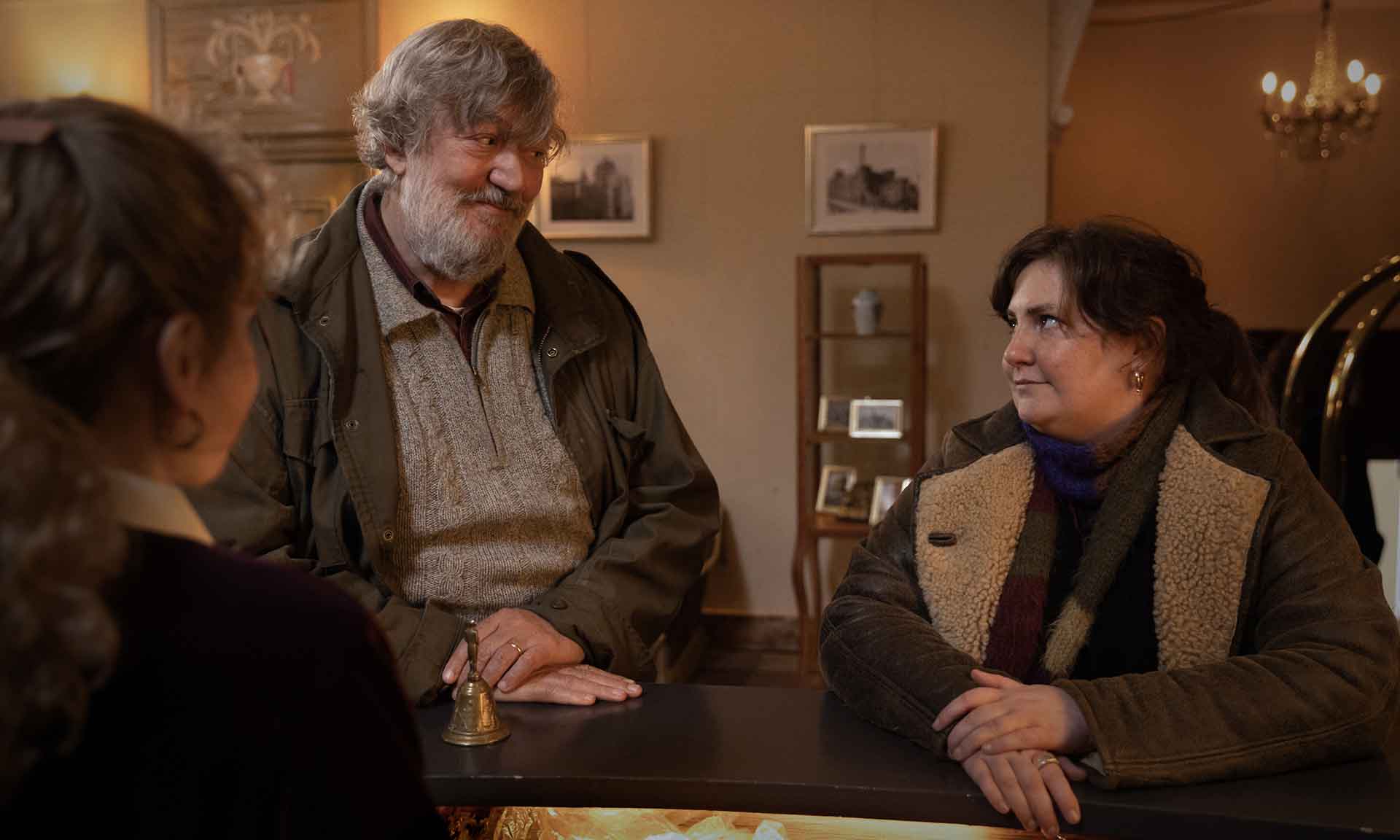 A tall man in a beard and a shorter woman in a large coat look at each other with a knowing look.