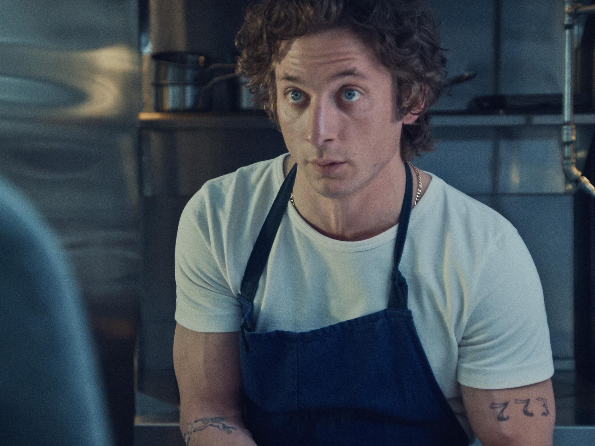 A white man in white t-shirt and black apron works in a kitchen, facing forward.