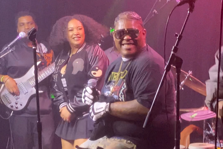 A man in a black t-shirt smiles, looking toward the camera in sunglasses, with a bassist and backup singer behind him