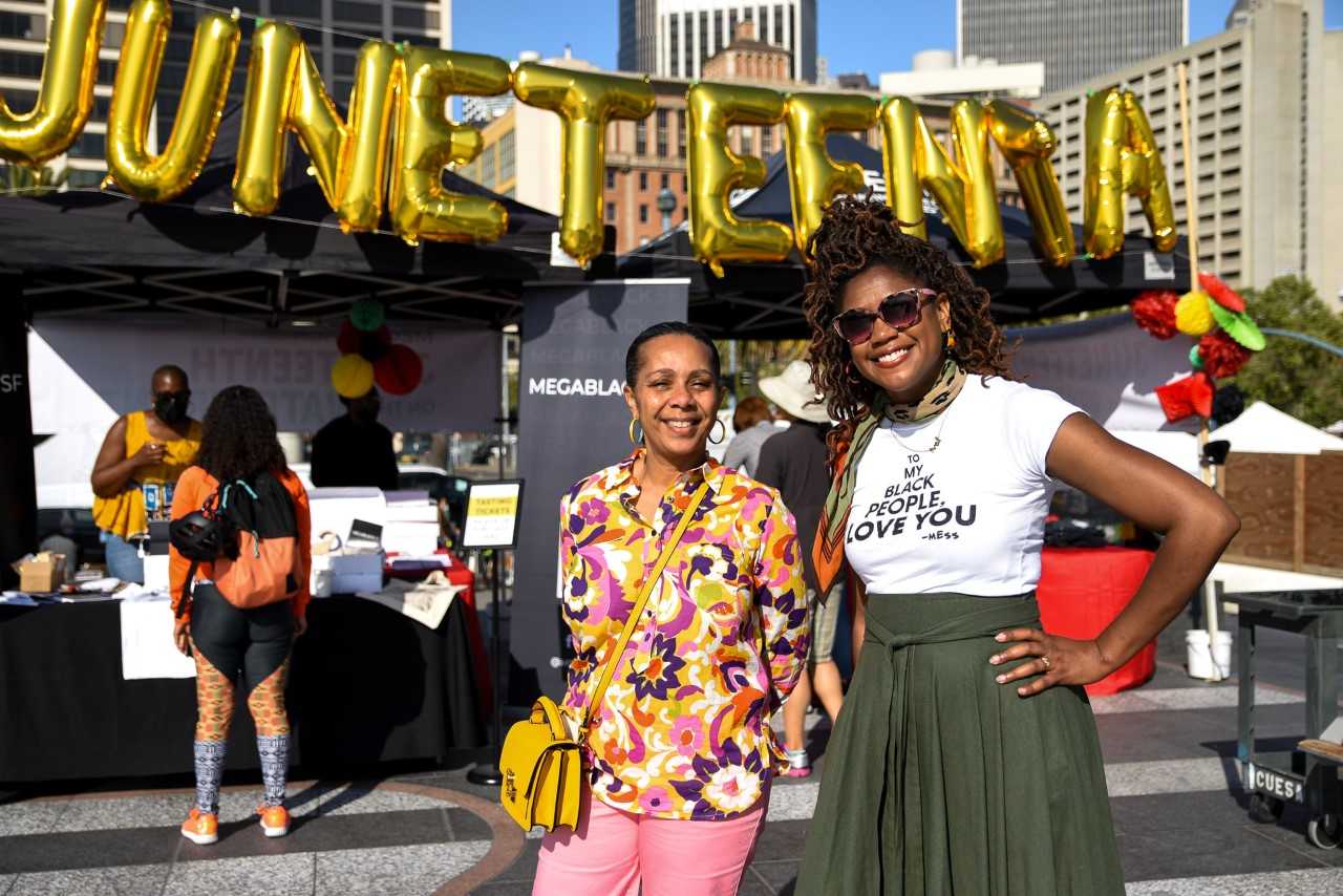 A set of balloons reads "JUNETEENTH" in golden lettering overhead, as two event attendees pose for a photo in the foreground.