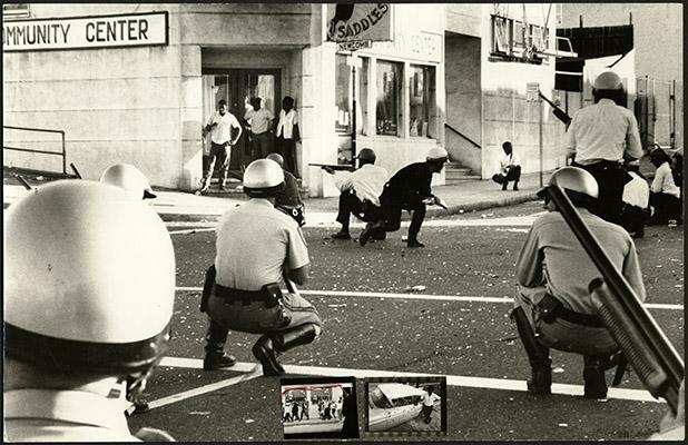 Black-and-white photograph of police crouched in street with guns while people stand in doorway of building 
