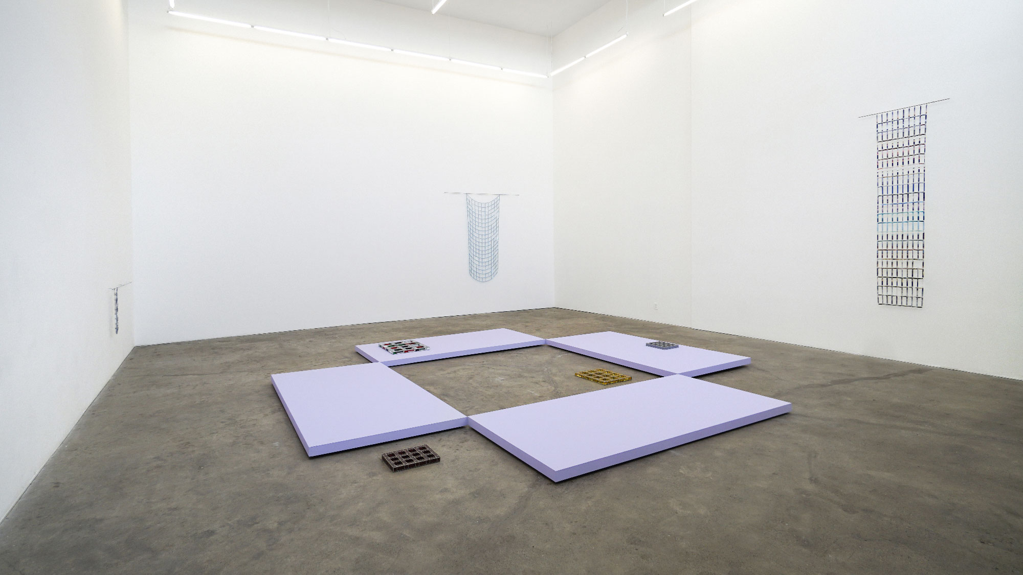 white-walled space with three minimal grid wall sculptures and large lavender platforms at center
