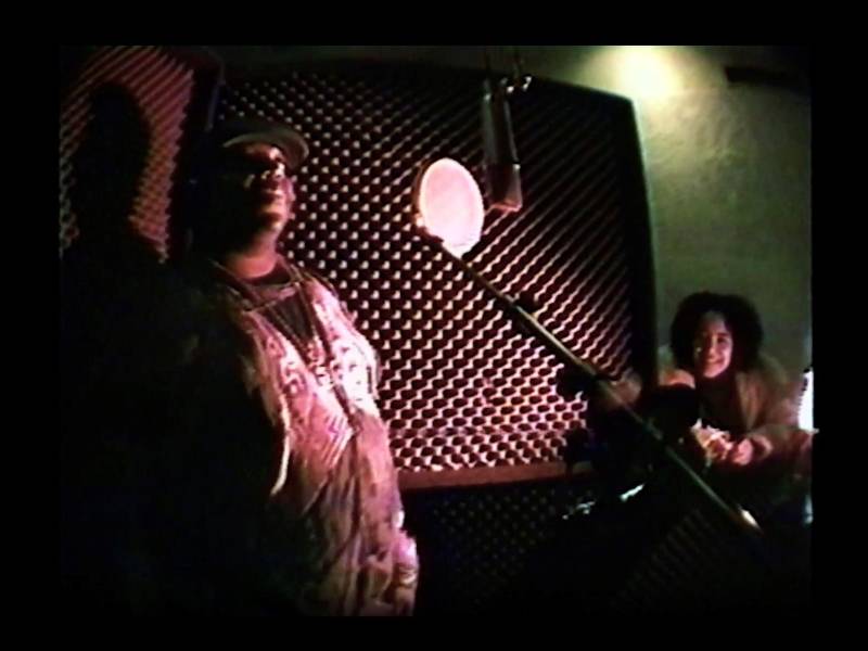 A large Black man stands behind a microphone in a studio while a smiling Black woman crouches nearby.