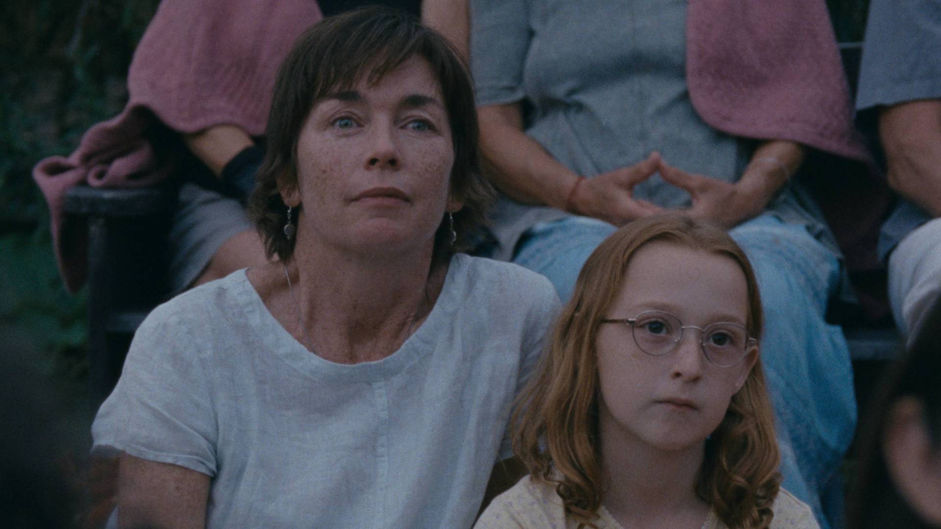 A middle-aged white woman sits with a young girl who is wearing glasses and looks bored.