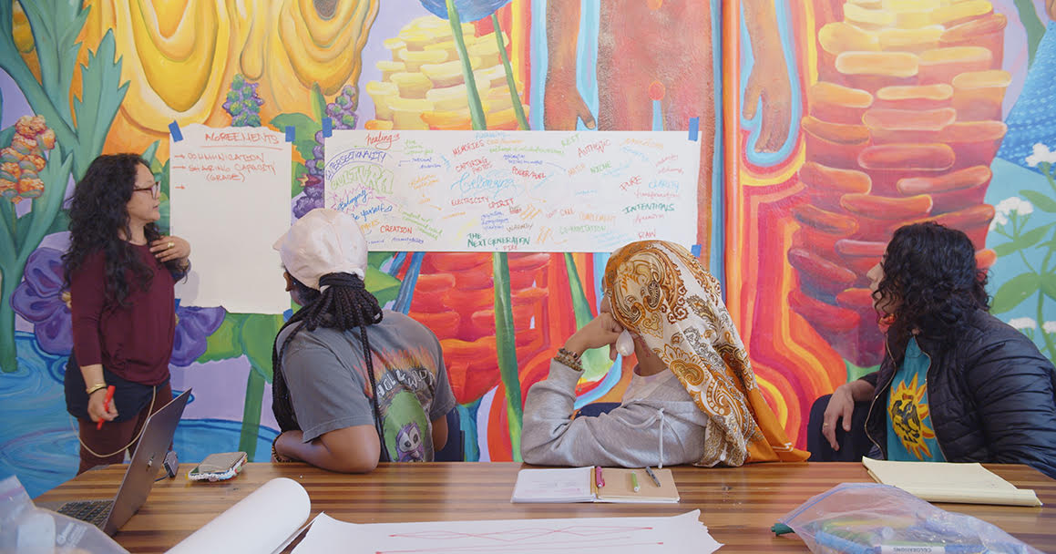 four people look at a sheet of paper with brainstorming ideas in a colorfully painted room
