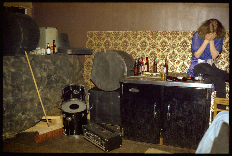A person sits with their head in their hands next to an array of beer bottles, drums and amps.