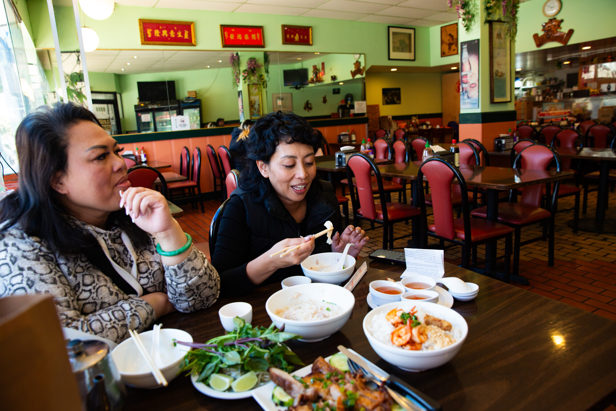 Two women eating noodles with a spread of Vietnamese food on the table in front of them.