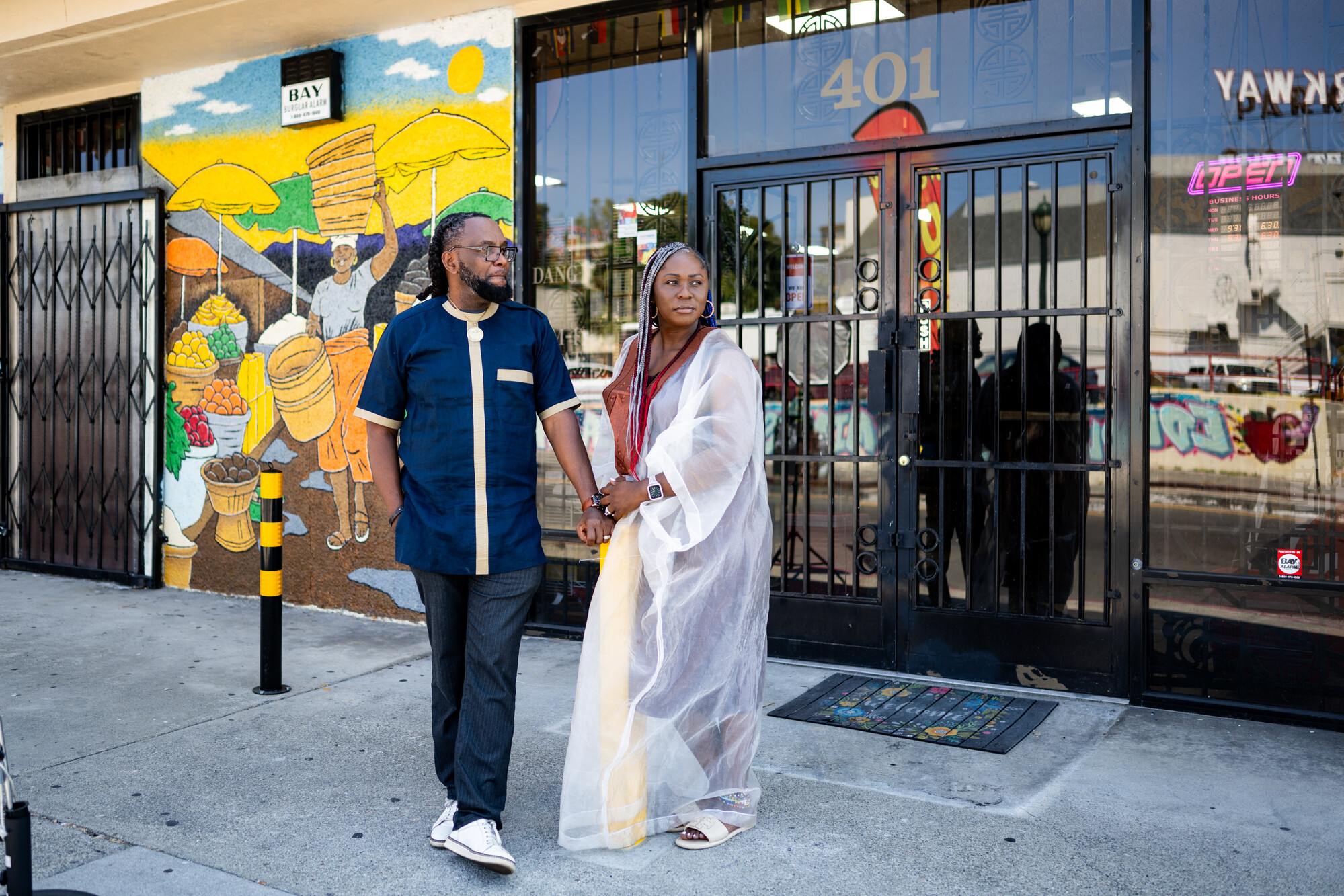 A man and woman stand in front of the yellow mural that decorates the front of their Afro-Caribbean market.