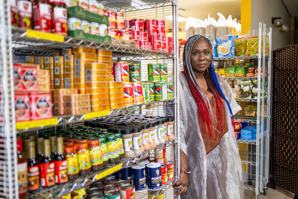 A woman with long red, white and blue braids poses next to a display of spices and tinned fish inside an African market.