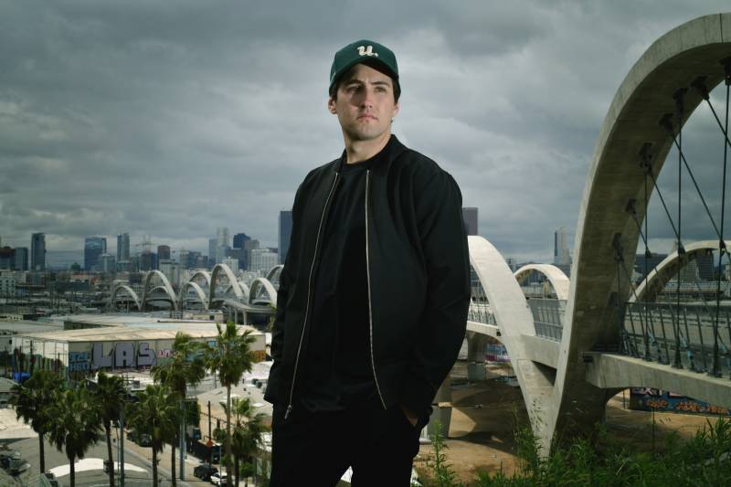 A white man in a baseball cap and black clothing stands in front of a cloudy sky and stares into the distance.