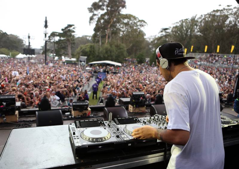 A male DJ in a white shirt and backwards baseball cap uses DJ equipment on a stage in front of a sea of people.