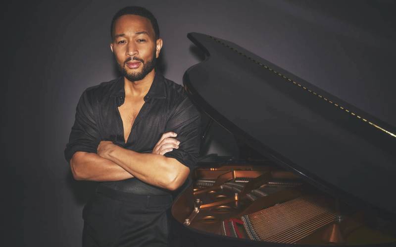 A fit Black man in a black button-down shirt opened at the top folds his arms while standing against a piano.