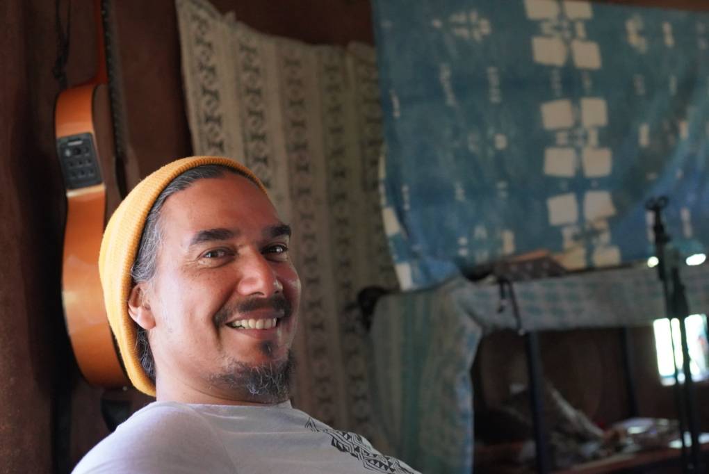 Ras K'dee smiles and looks at the camera, while wearing a yellow knitted cap and sitting in his music studio.
