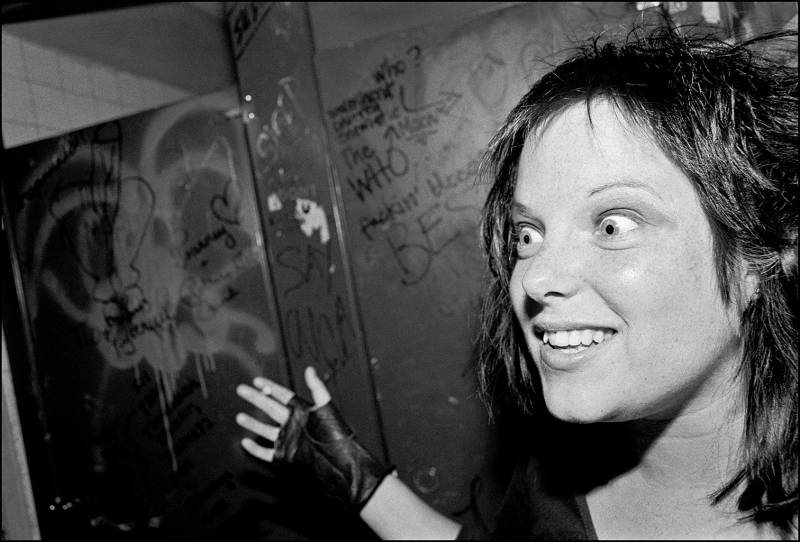 A wild-eyed young woman wearing fingerless gloves smiles in a dank, graffitied room.