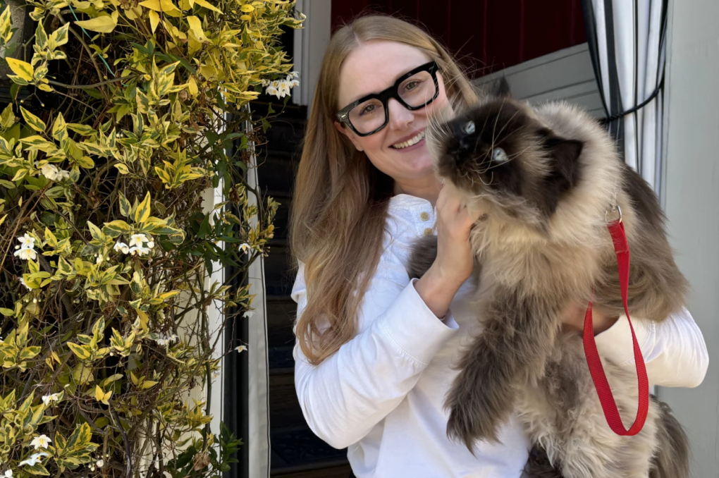 A white woman with long fair hair and eye glasses holds a fluffy cat that's wearing a harness.