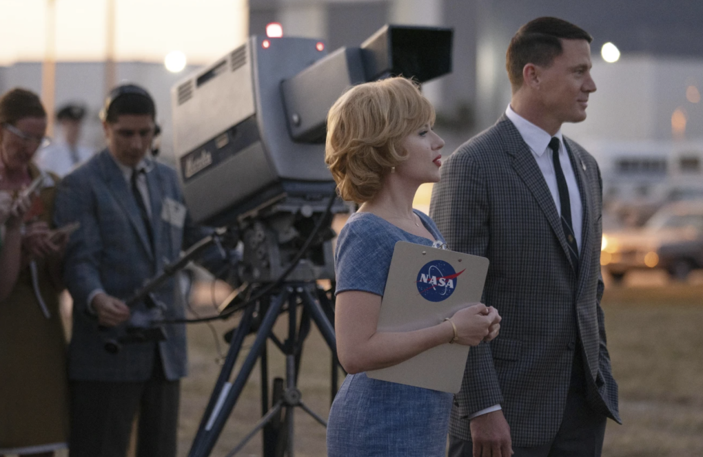 A white woman and white man stand outdoors with a large television camera behind them. She is holding a NASA clipboard.