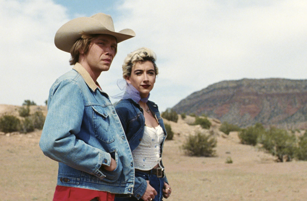 A young man wearing blue denim and a cowboy hat stands out on a dirt road with a gender non-conforming person wearing short bleached hair and striking makeup.