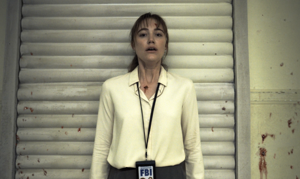 A stunned white woman wearing a blouse and FBI badge stands in front of a blood-spattered wall.