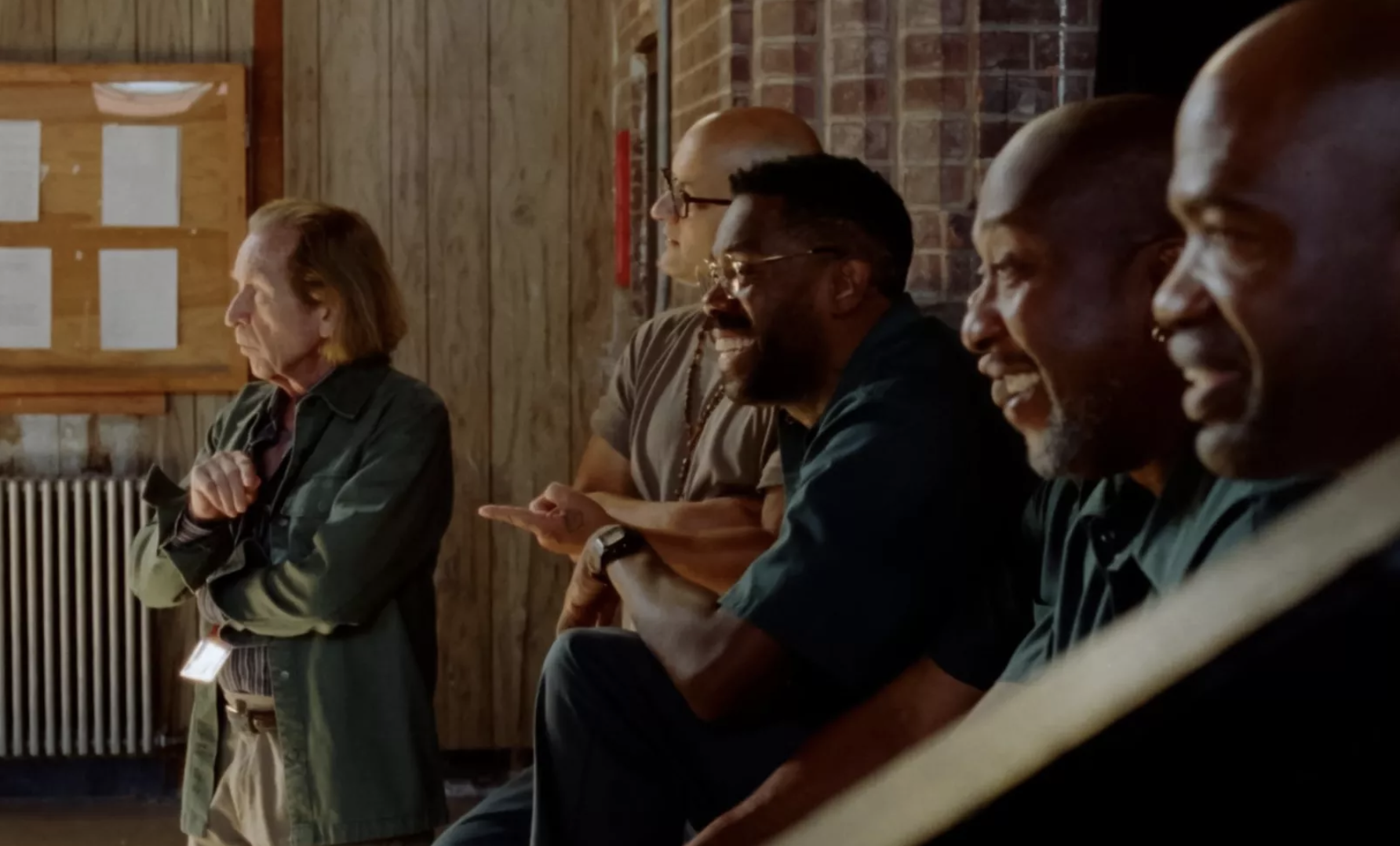 Five men gather at the back of the room, each watching something off-camera. Three of the men are laughing. 