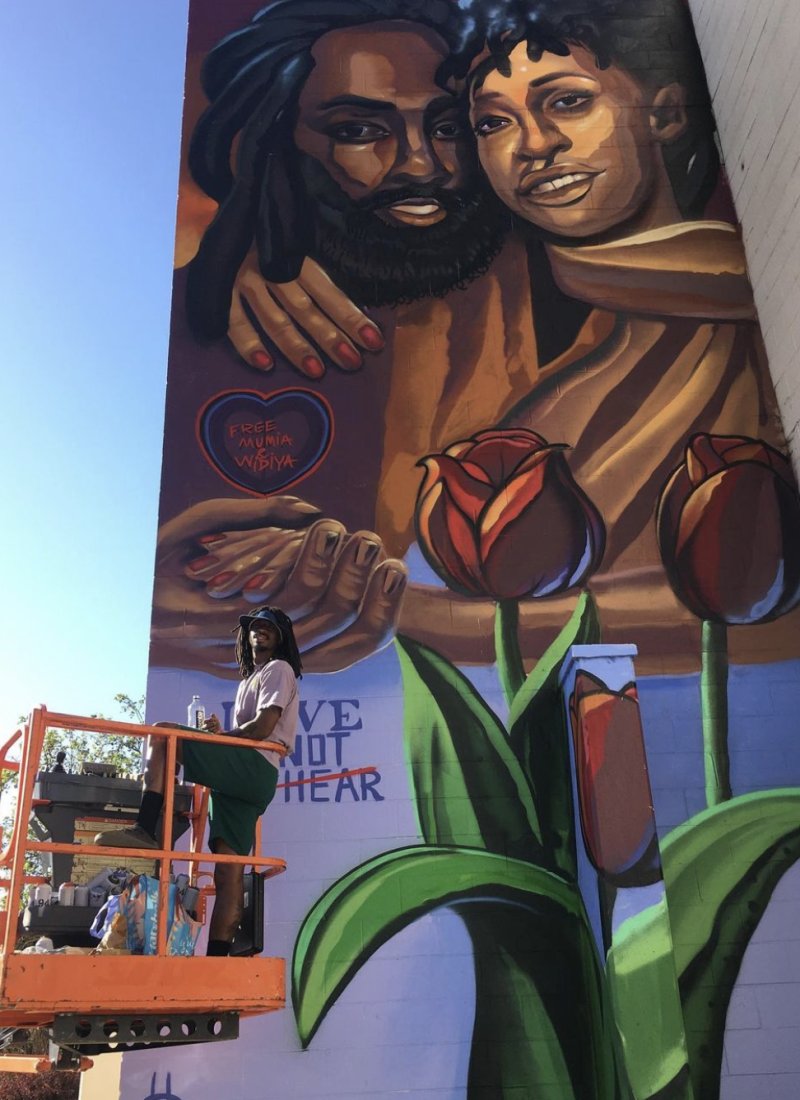 Timothy B. stands on a lift in front of a mural he painted at the East Oakland Youth Center dedicated to journalist and political prisoner Mumia Abu-Jamal.
