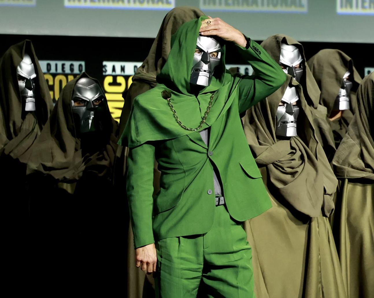 A man in a striking green suit and metal mask stands with a group of other people wearing metal masks and robes.