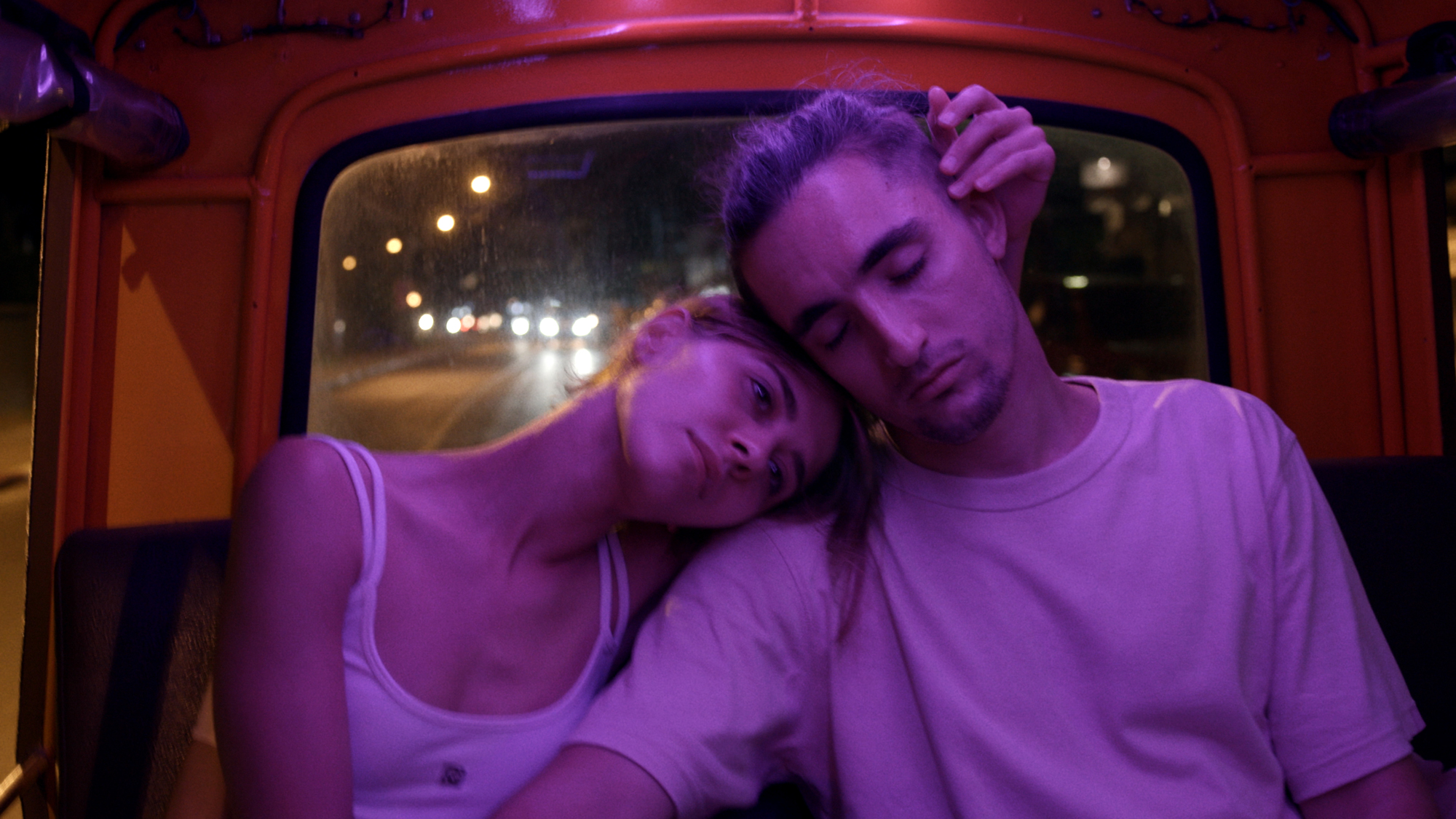 Two people lean on each other in back seat of car under purple light